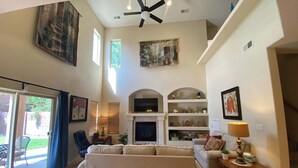 A spacious living room with a large sofa, a television, and a fireplace. There are multiple shelves with decorative items and a ceiling fan. - Amazing 4-bedroom: AC, WiFi, 2 car garage with river view in NW Albuquerque (Albuquerque)