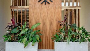 Zona de recepción con un panel de madera que muestra el nombre "CASAVANGLAR" y un diseño moderno y geométrico. Hay dos grandes macetas con plantas verdes.