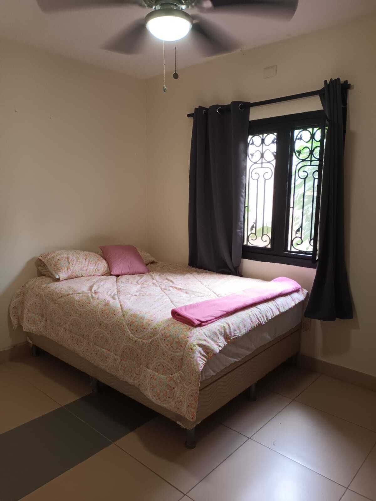 Quarto com cama, ventilador de teto, janela com cortinas e vista para o verde lá fora.