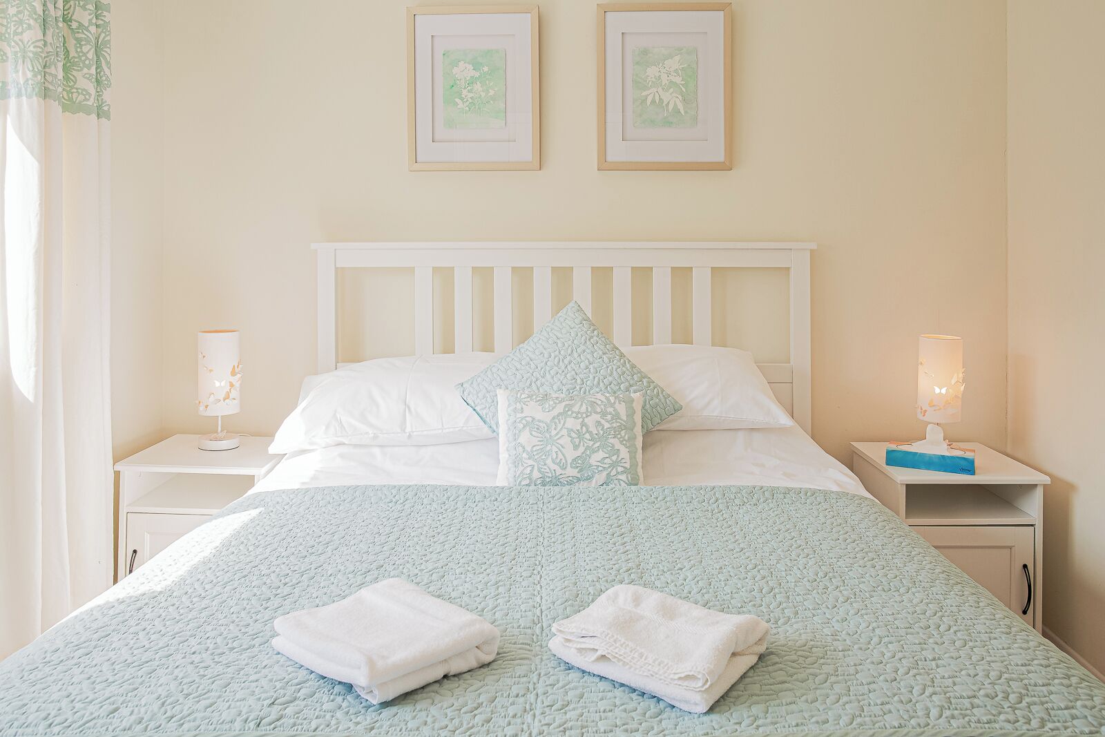 A neatly made bed with a white headboard, two pillows, and a blue quilt. There are two bedside tables with lamps and a framed picture on the wall above the bed.