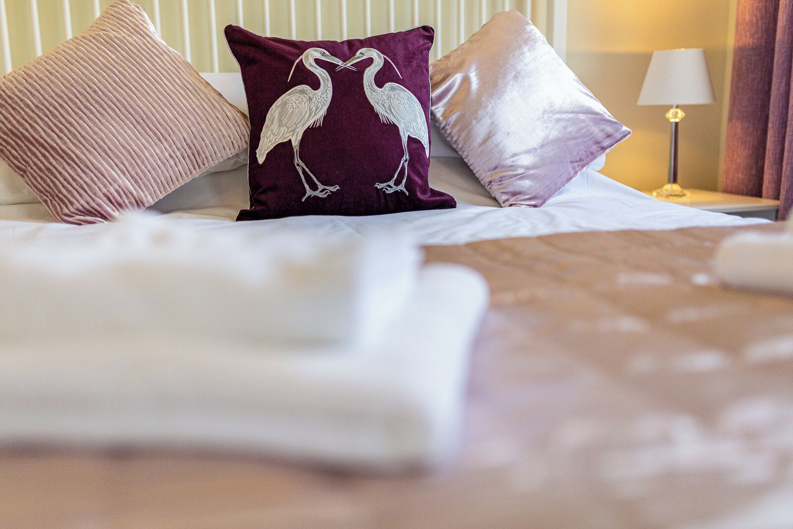 Un lit bien fait, avec du linge de lit blanc, un coussin violet orné d’un motif d’oiseau et un coussin rose.