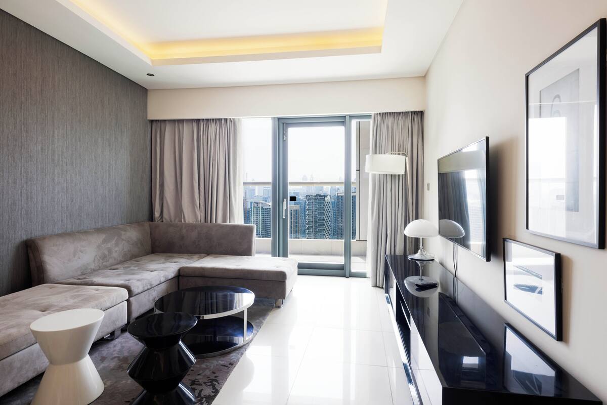 A modern hotel room with a sofa, coffee table, TV, and large windows offering a city view.