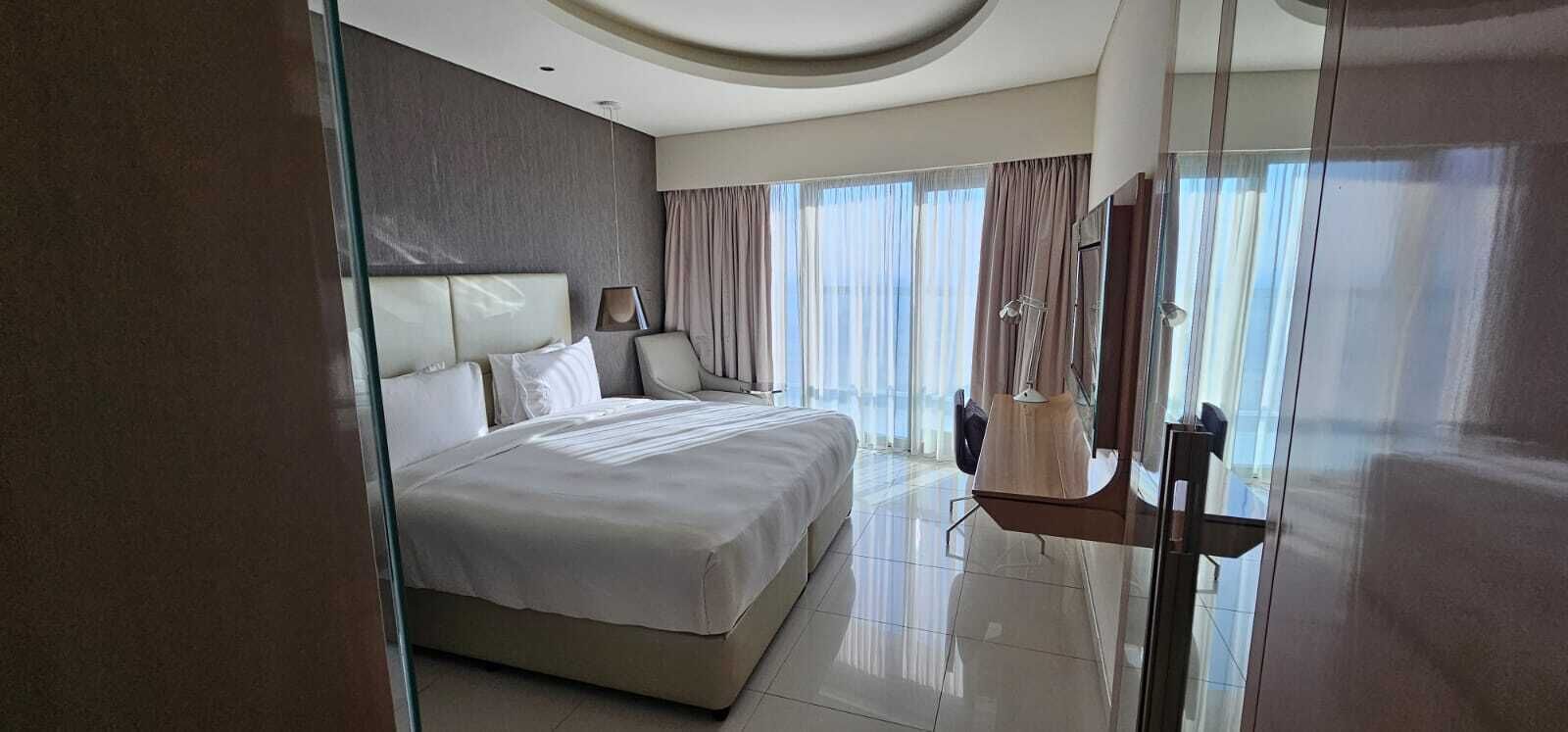 Une chambre d’hôtel moderne dotée d’un grand lit, d’une cloison en verre, d’un coin salon avec un fauteuil et d’une grande fenêtre avec des rideaux.