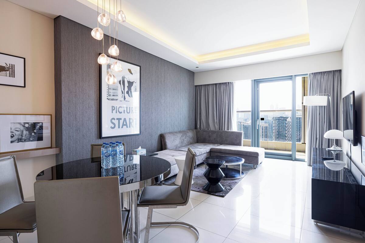 A modern hotel room with a dining area, a sofa, a TV, and a large window with a city view.