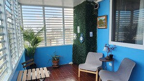 Une pièce avec un mur bleu, un ventilateur de plafond, une fenêtre avec des stores, une décoration murale verte, un banc en bois, une chaise grise et une petite table avec une lampe.