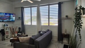 Un salon avec un canapé, un téléviseur fixé au mur, une fenêtre avec des stores et une plante.