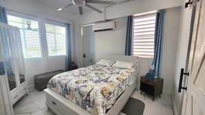 Une chambre à coucher comprenant un lit, un ventilateur de plafond, un appareil de climatisation, une table de chevet avec une lampe et une fenêtre avec des rideaux.