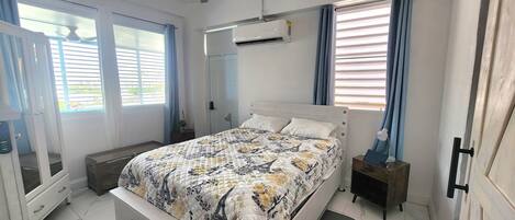 A bedroom with a bed, a ceiling fan, an air conditioning unit, a bedside table with a lamp, and a window with curtains.