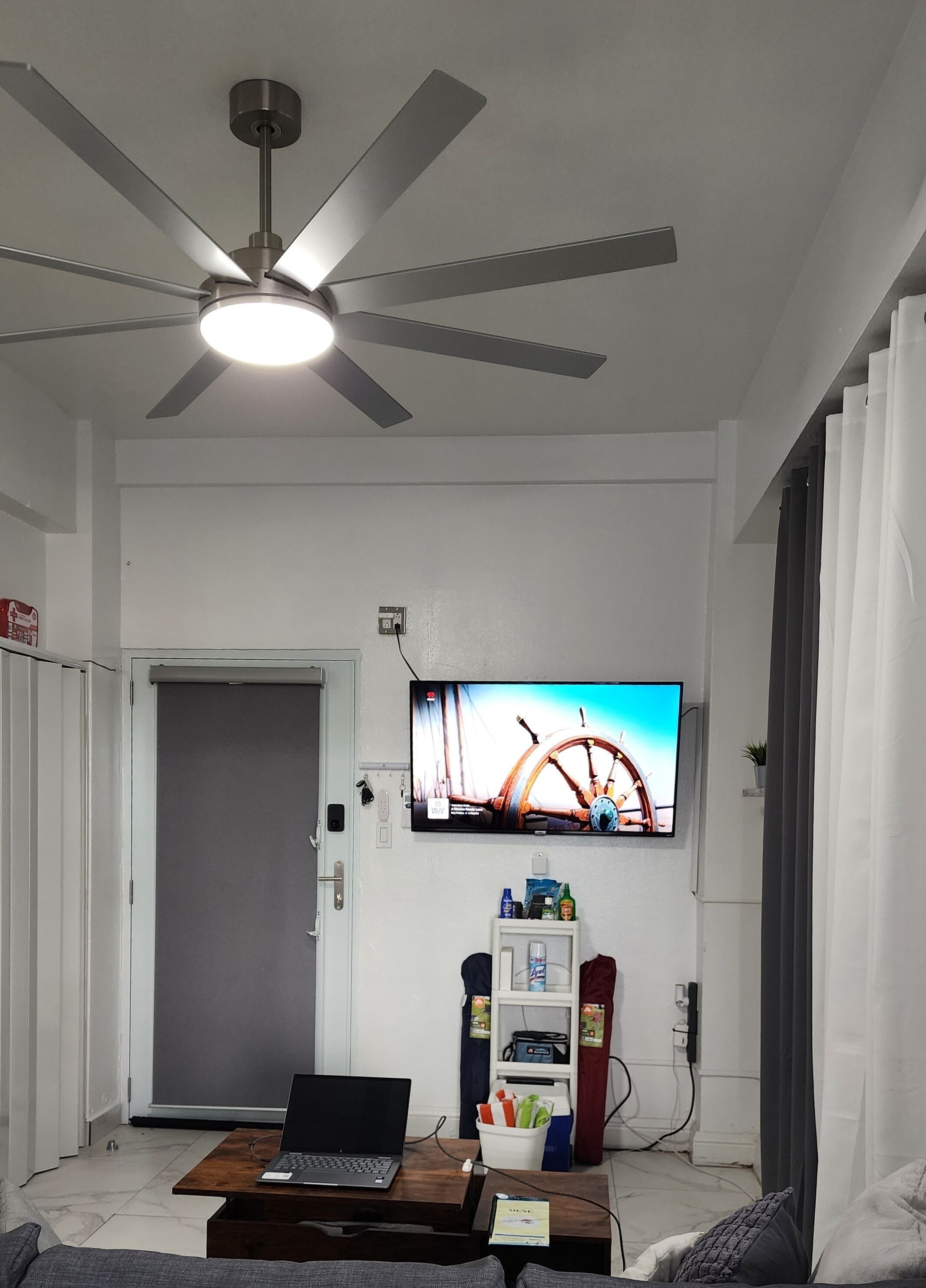 Una sala de estar con ventilador de techo, un televisor montado en la pared, una computadora portátil sobre una mesa de centro y una puerta que da a otra habitación.