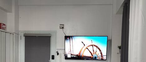 Una sala de estar con ventilador de techo, un televisor montado en la pared, una computadora portátil sobre una mesa de centro y una puerta que da a otra habitación.