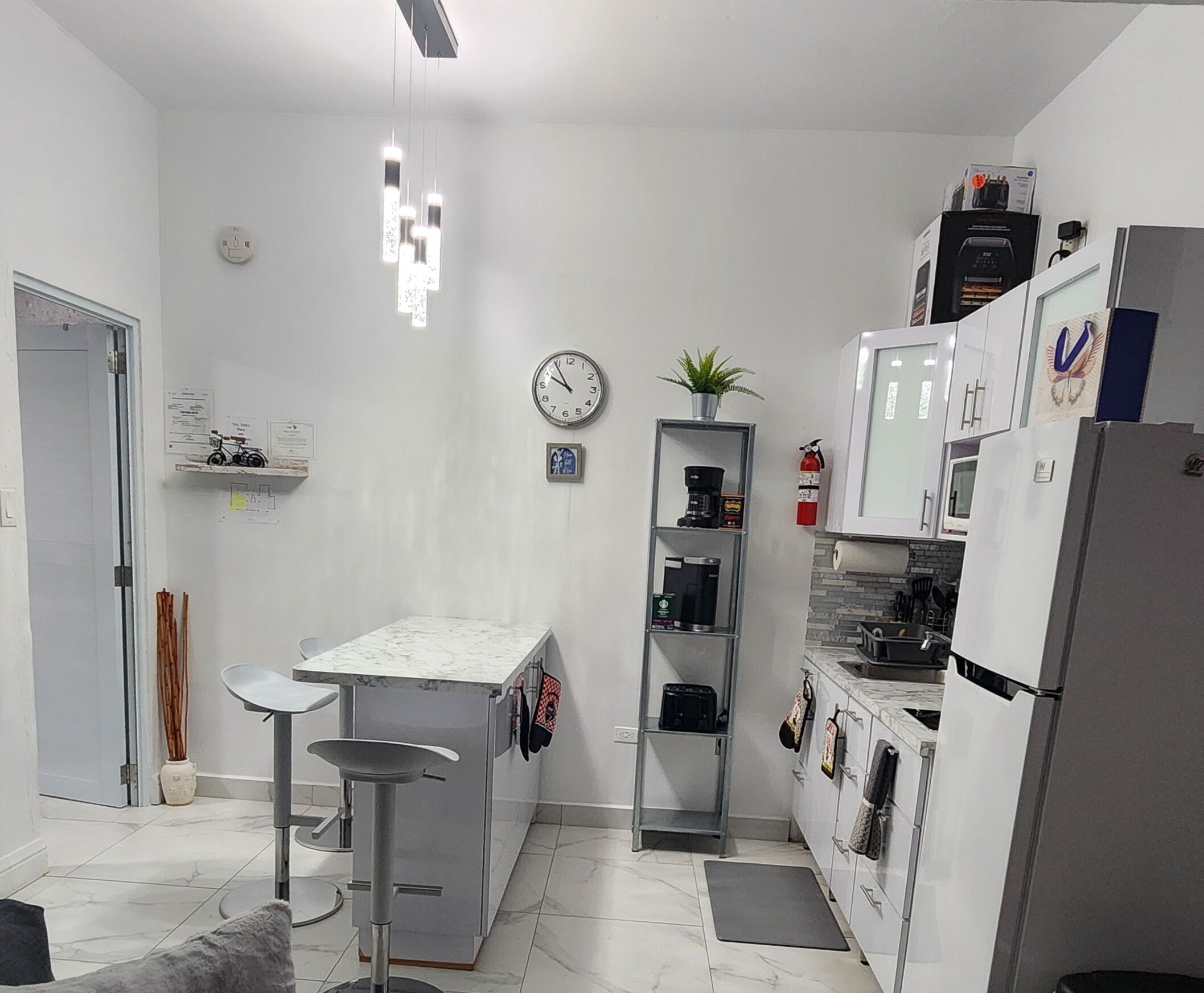 Una cocina moderna con armarios blancos, encimera de mármol, una cafetera, un reloj y una pequeña planta.