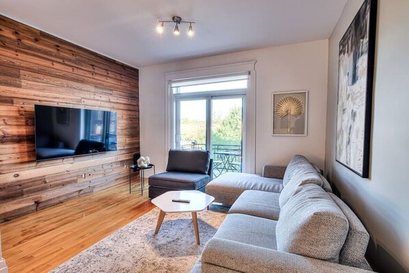 A modern living room with a flat-screen TV mounted on a wooden wall, a grey sofa, a small table, and a window with a view of greenery. - Spacious 3BR in Downtown Montreal (Montréal)