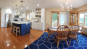 A modern kitchen with white cabinets, a central island, and a dining area with wooden chairs and a blue rug.