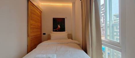 A small, neatly made bed with white linens and a folded towel, a wooden wardrobe, a painting of a deer, and a window with a view of buildings.