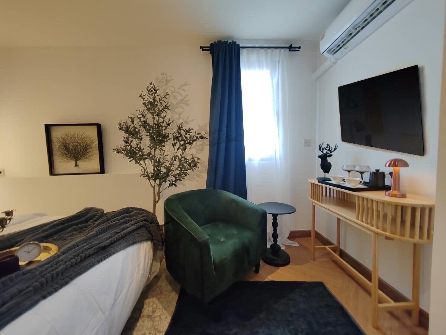 A hotel room with a bed, a green armchair, a small table, a TV, a plant, and a window with curtains.