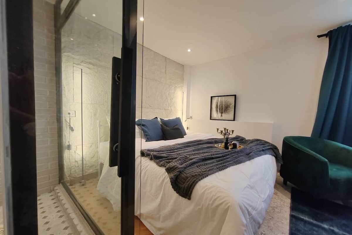 A modern bedroom with a glass shower enclosure, a bed with white linens, a dark green chair, and a framed artwork on the wall.