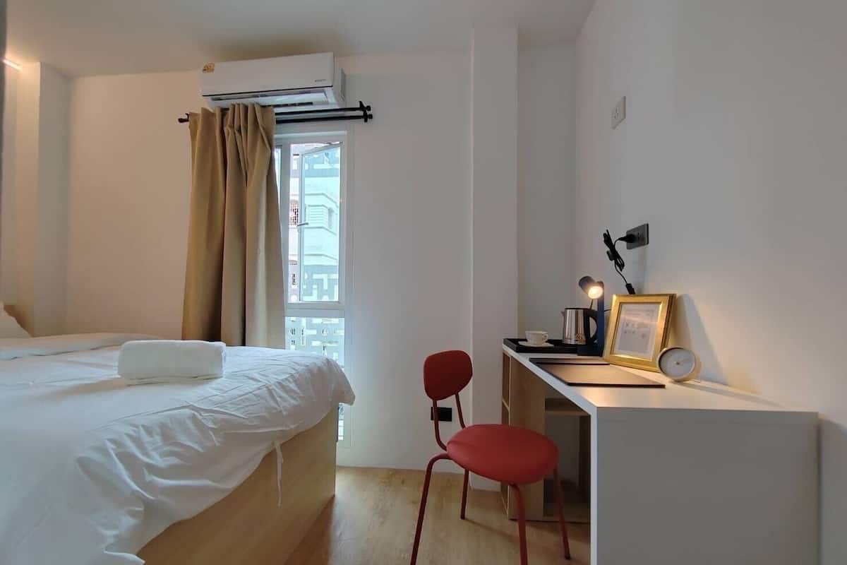 A hotel room with a bed, a desk with a chair, a small table with a kettle, a picture frame, and an air conditioning unit.