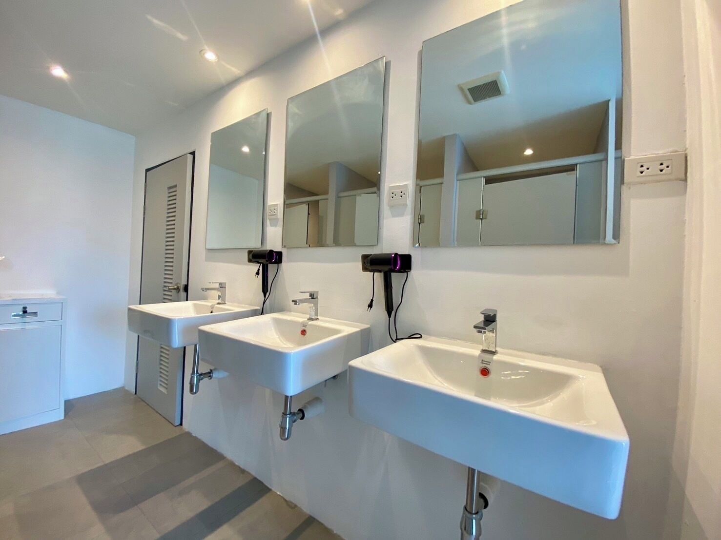 A modern bathroom with three white sinks, each with a mirror above. There are hair dryers mounted on the wall, and a small cabinet is visible to the left.