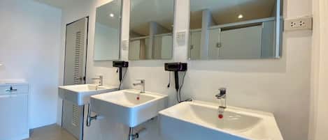A modern bathroom with three white sinks, each with a mirror above. There are hair dryers mounted on the wall, and a small cabinet is visible to the left.