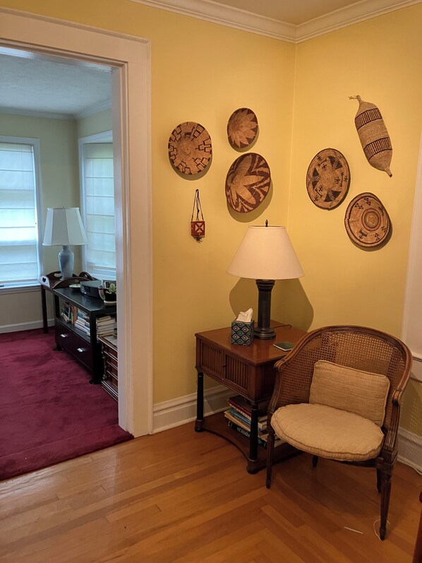 A room with a wooden floor, a wicker chair, a small table with a lamp, and wall decorations.