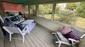 Ein überdachter Außenbereich mit Grill, zwei weißen Stühlen, einem Tisch und einer Bank mit kariertem Sitzkissen.