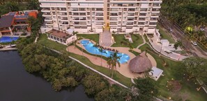 Un complejo turístico con una piscina grande, una estructura con techo de paja y un edificio de varios pisos con balcones.