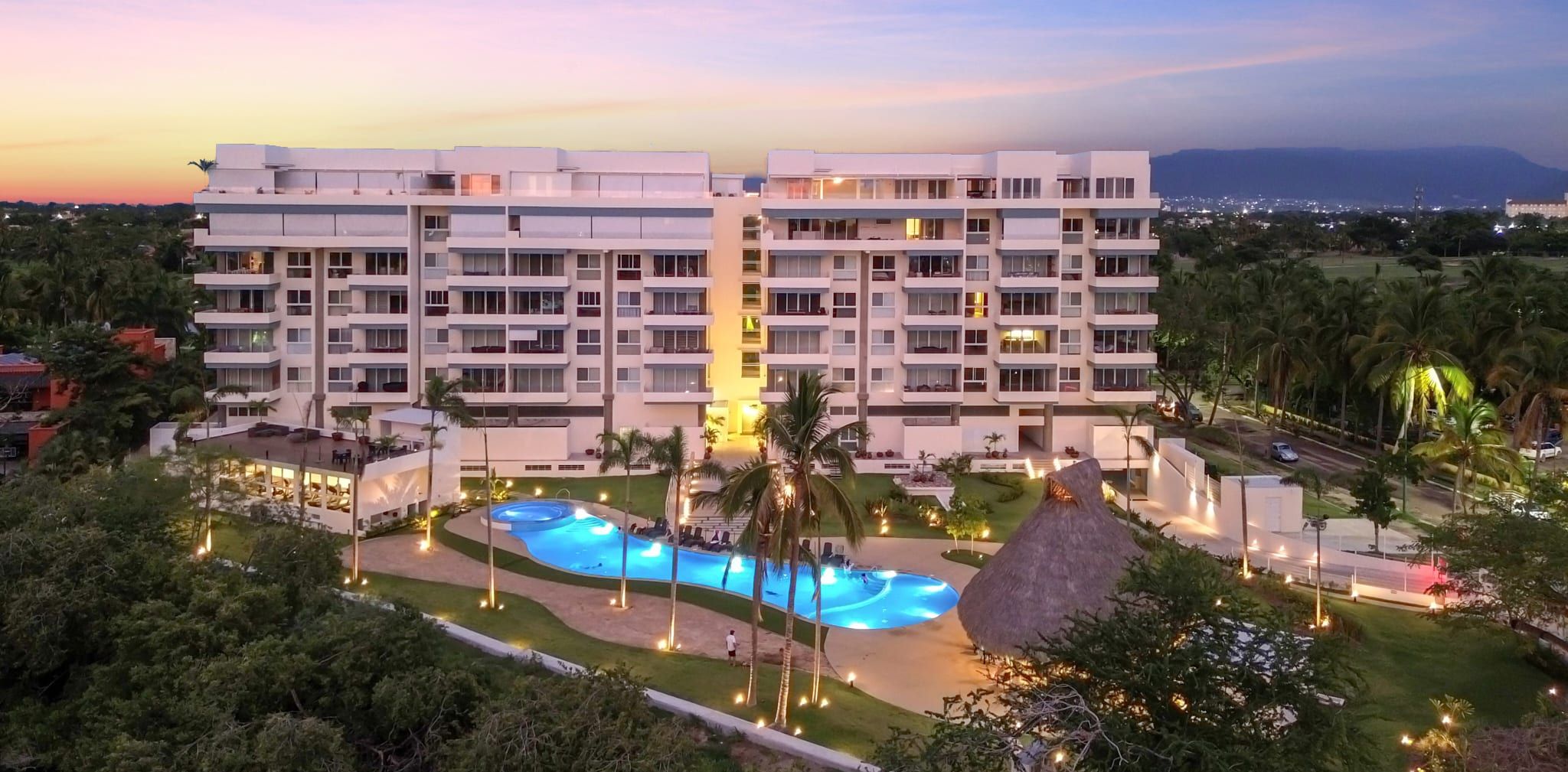 Ein mehrstöckiges Hotel mit Swimmingpool, umgeben von Palmen und Beleuchtung.