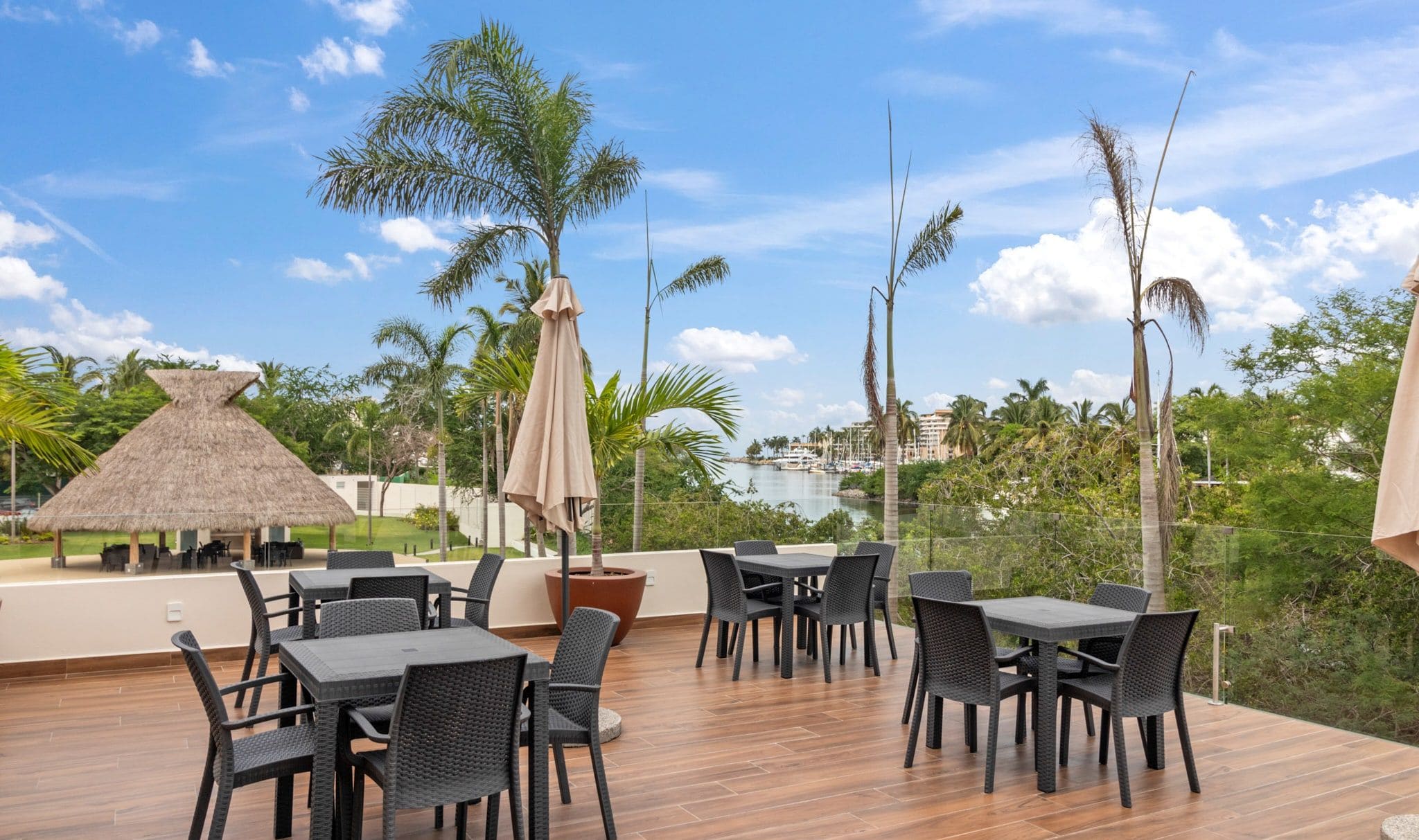 An outdoor dining area with tables and chairs, a thatched-roof structure, and a view of a marina and buildings in the distance.