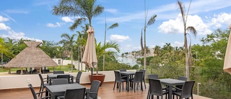 Un área de comedor al aire libre con mesas y sillas, una estructura de techo de paja y vistas a un puerto deportivo y edificios a lo lejos.