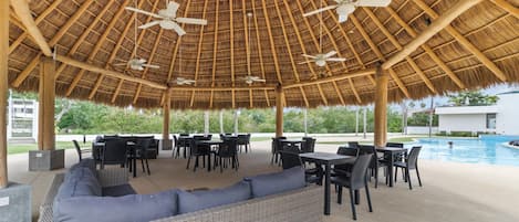 An outdoor seating area with tables and chairs, a pool, and a thatched roof structure.