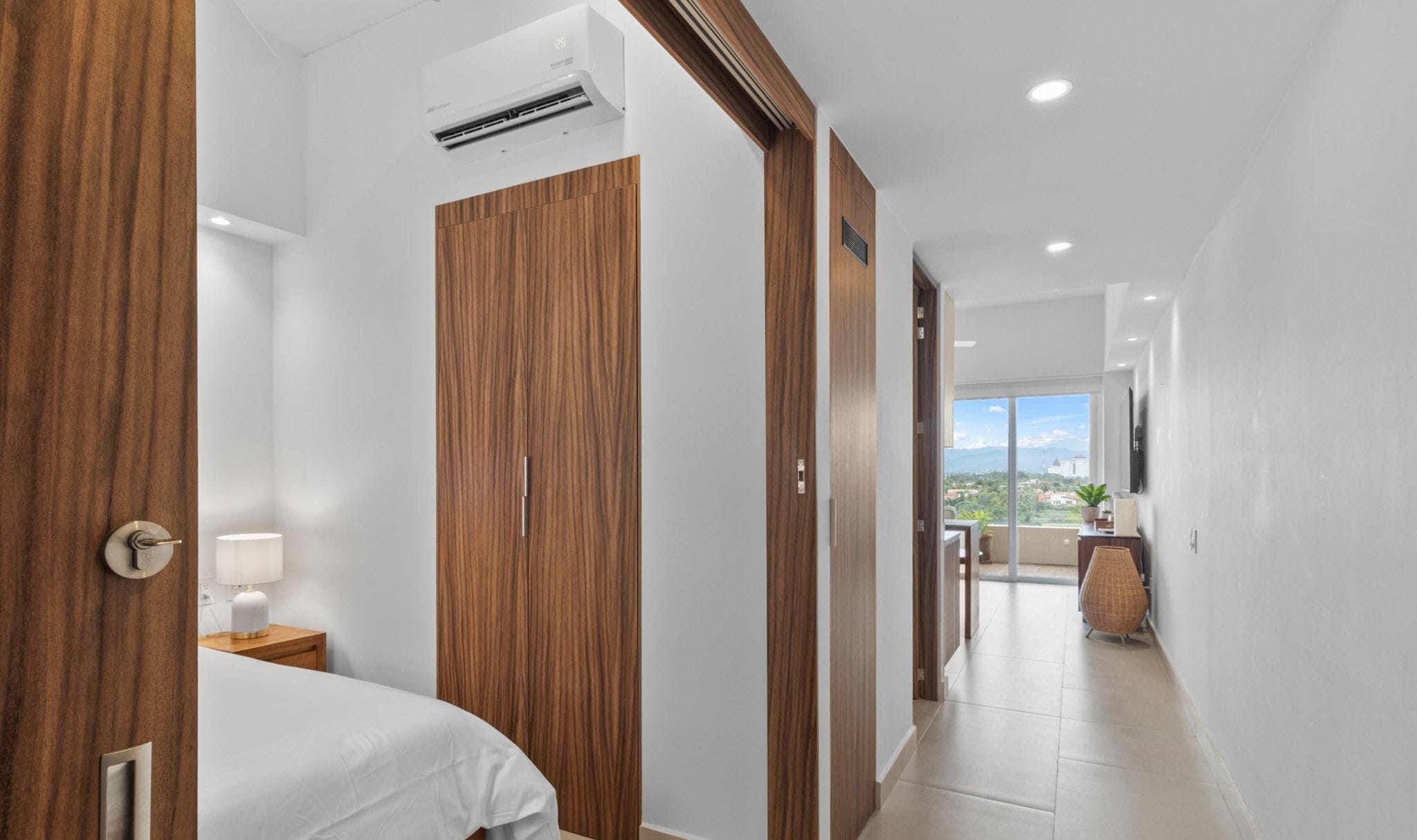 Pasillo de hotel con puertas de madera, una cama, una mesita de noche y vistas al exterior.