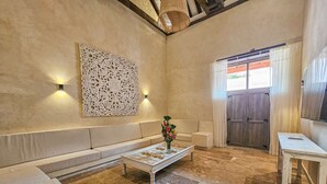 Un ampio soggiorno con un divano beige, un tavolino da caffè con un centrotavola floreale, un elemento decorativo a parete e una porta in legno con tende.