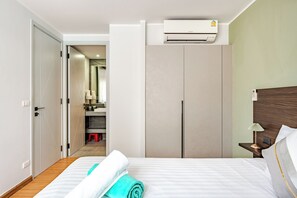 A hotel room with a bed, a nightstand, a bedside table, an air conditioning unit, a wardrobe, and a bathroom visible through an open door. - Modern 61m² Apt - Central Chalong/ Fast WiFi (Phuket)