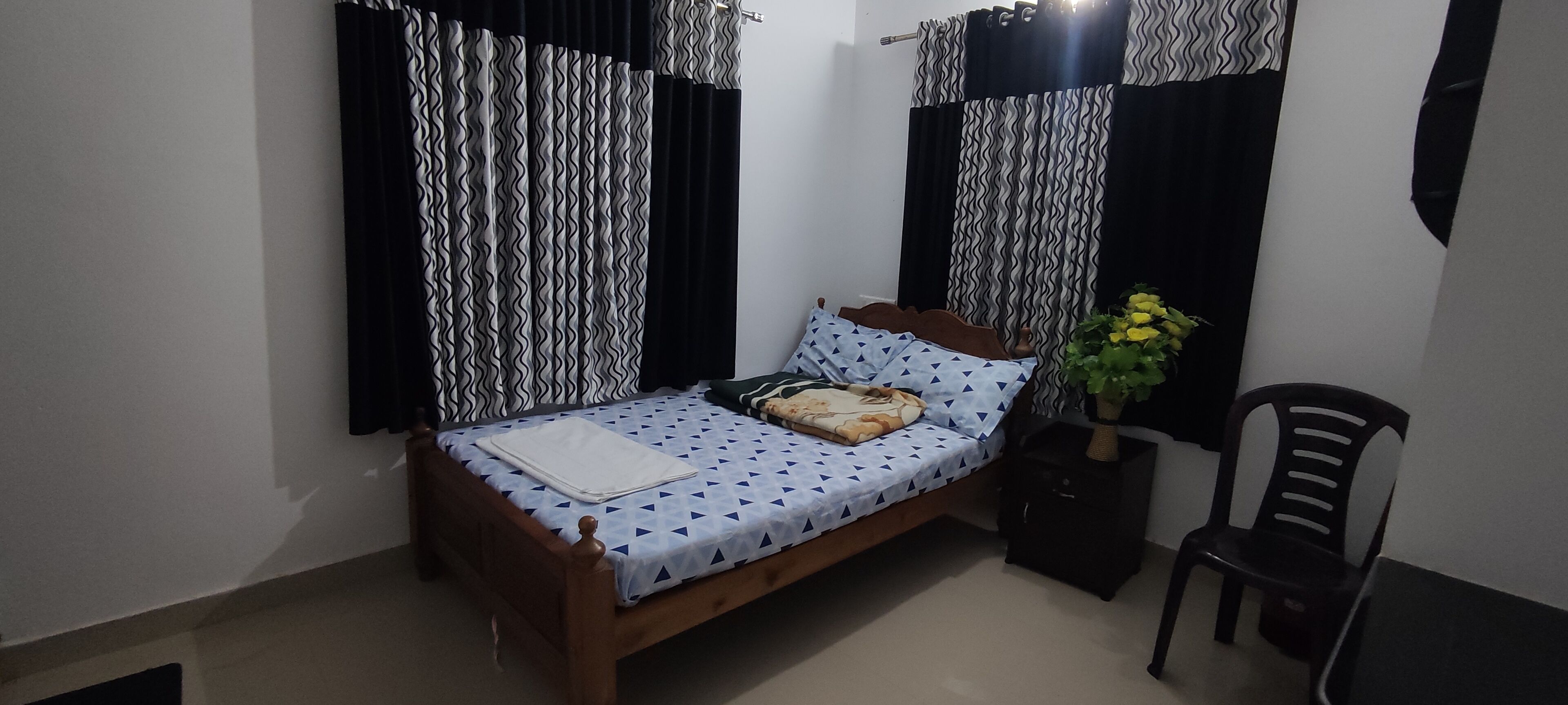A bedroom with a wooden bed, patterned bedding, black and white curtains, a small table with a plant, and a chair.