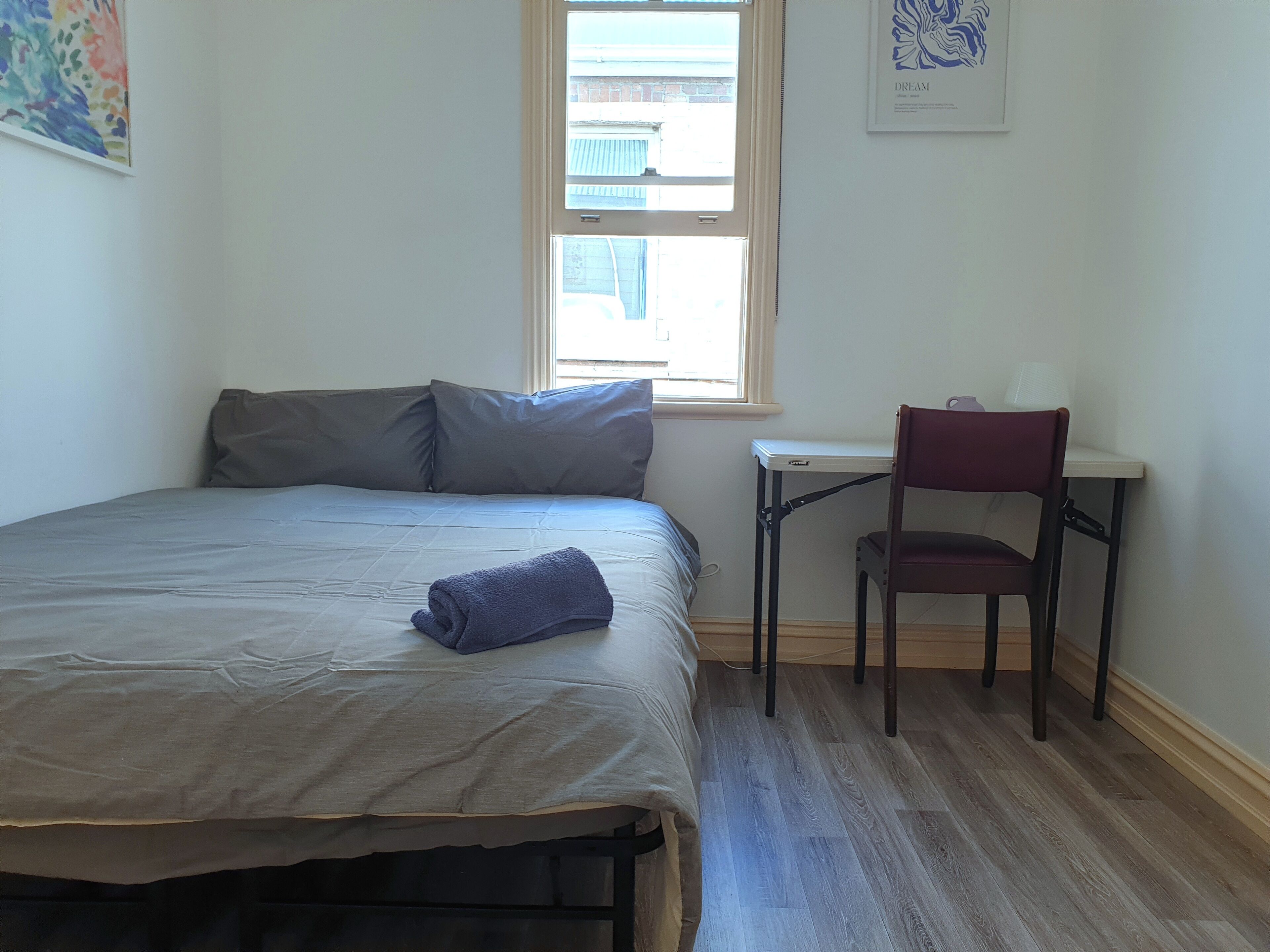 A neatly made bed with pillows, a small desk with a chair, and a window with a view of buildings.