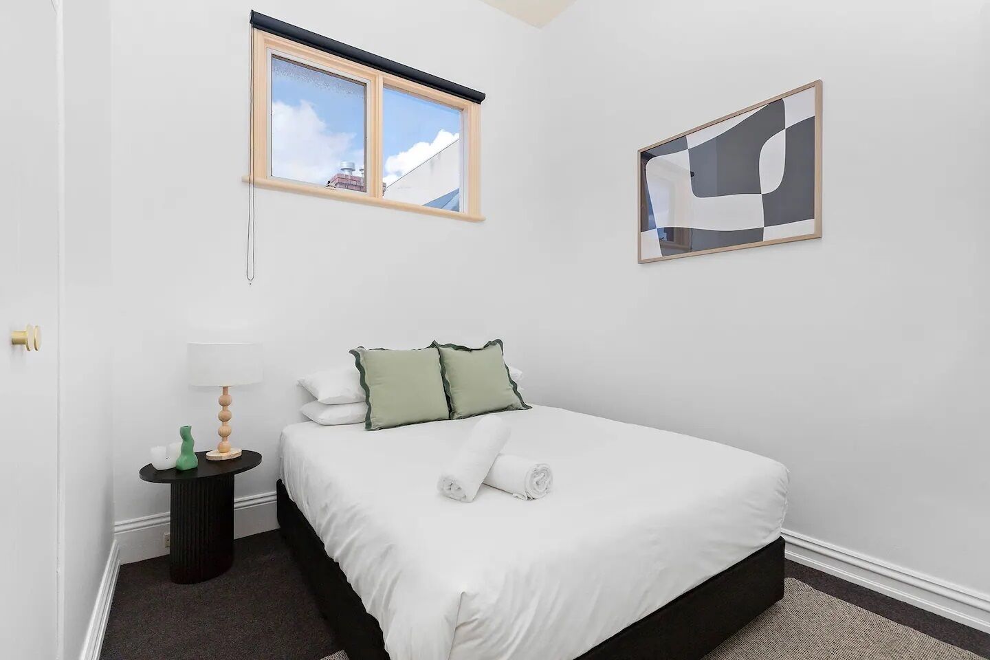 Een keurig opgemaakt slaapkamer met een bed, nachtkastje, lamp, raam en een abstract kunstwerk aan de muur.