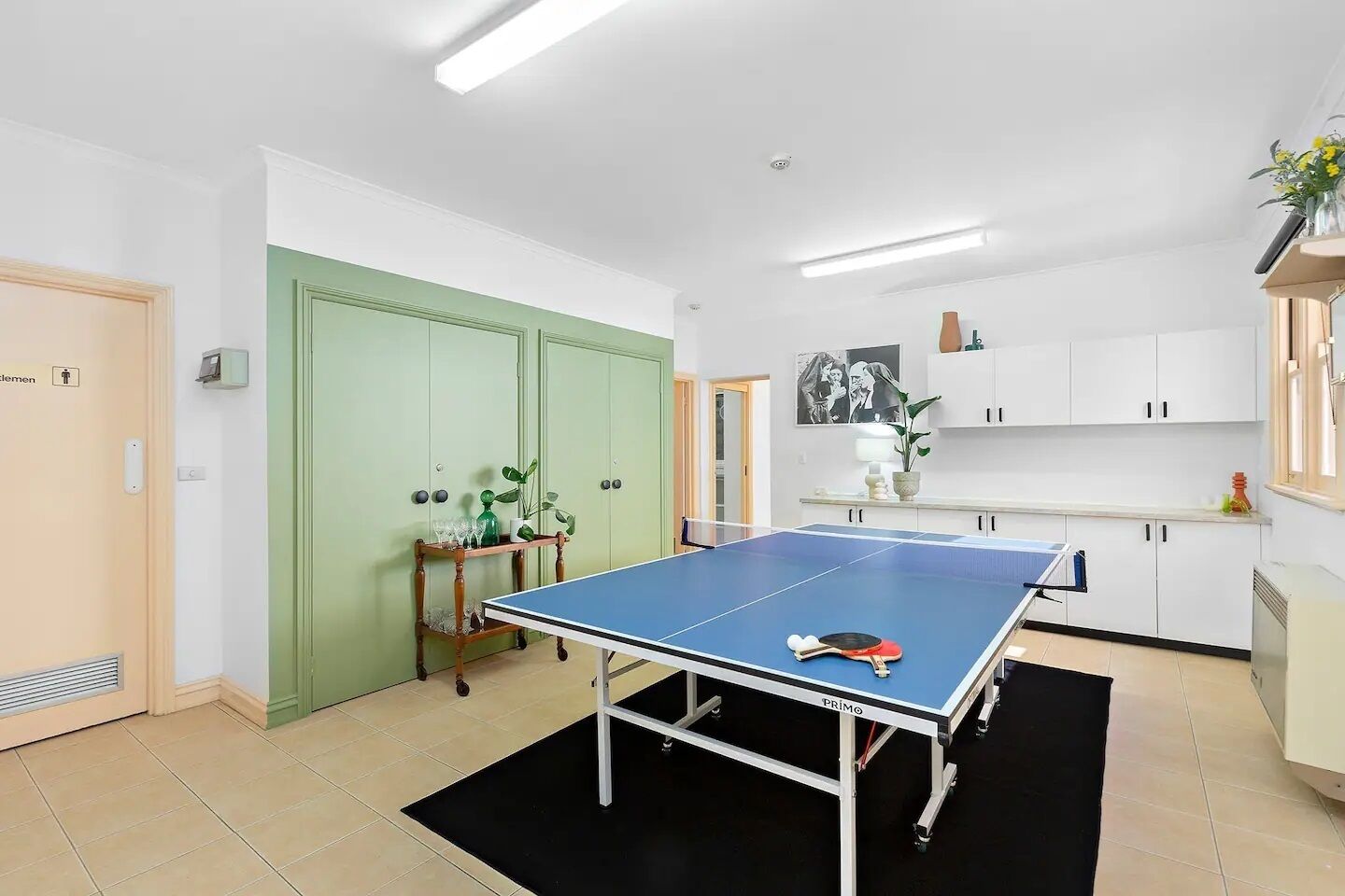Ein Zimmer mit Tischtennisplatte, einer grünen Tür, einer kleinen Pflanze und einer Küchenzeile mit weißen Schränken.