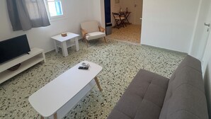 A living room with a sofa, a coffee table, a TV, and a small table with a chair. There is a window with curtains, an air conditioning unit, and a hanging light fixture. - Selini KHORA (Mikonos)