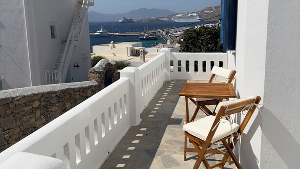 A balcony with a stone floor, white walls, and a view of the sea and boats. - Selini KHORA (Mikonos)