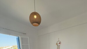 A bedroom with a bed, bedside lamps, a hanging light fixture, a window, and a wall decoration. - Selini KHORA (Mikonos)