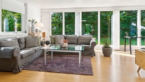 Una sala de estar moderna con un sofá gris, una mesa de centro de cristal y vistas a un jardín exuberante a través de grandes ventanales.