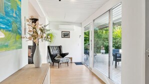 Un interior moderno con un banco de madera, un sillón de cuero negro, un jarrón blanco con plantas y un amplio ventanal que da a un patio exterior.