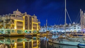 A marina with boats docked, a building with arched windows, and a reflection in the water.