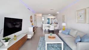 A modern living room with a flat-screen TV, white sofas, a dining area, and a kitchen in the background.
