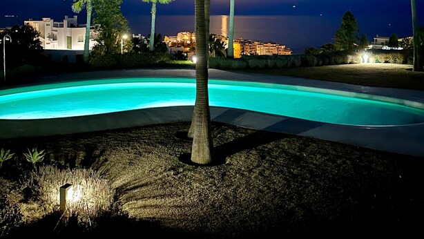 A nighttime scene with a swimming pool, palm trees, and a cityscape in the background.