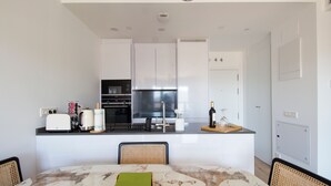 Eine moderne Küche mit weißen Schränken, einem schwarzen Kühlschrank und einer Marmorarbeitsplatte. Ein Esstisch mit Stühlen und einer grünen Tischdecke ist ebenfalls vorhanden.
