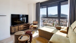 Ein modernes Wohnzimmer mit großem Fernseher, gemütlicher Sitzgelegenheit und Blick auf eine Stadtlandschaft durch große Fenster.