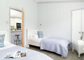 Une chambre avec deux lits, un petit tabouret, une chaise en osier et une porte donnant sur la cuisine.