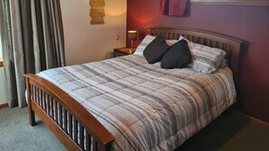 A neatly made bed with a striped comforter and two black pillows, a wooden headboard, a nightstand with a lamp, and a wall with a 'Love and Joy' sign. - Home Sweet Home (Timaru)
