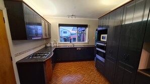 A compact kitchen with dark cabinets, a herringbone wood floor, a window with a view of houses, and a built-in microwave. - Home Sweet Home (Timaru)